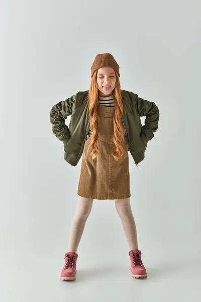 Menina com cabelos longos e chapéu de inverno na cabeça colando língua e de pé em vestido e jaqueta em cinza — Fotografia de Stock