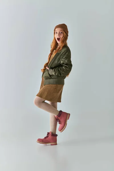 Зимняя мода, изумленная девушка с длинными волосами и вязаной шляпой, стоящая в платье и куртке на сером — стоковое фото