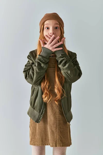 Зимняя мода, шокированная девушка с длинными волосами и вязаной шляпой, стоящая в платье и куртке на сером — стоковое фото