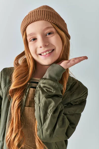 Зимняя мода, счастливая девушка с длинными волосами и вязаной шляпой стоя в пиджаке цвета хаки на сером фоне — стоковое фото