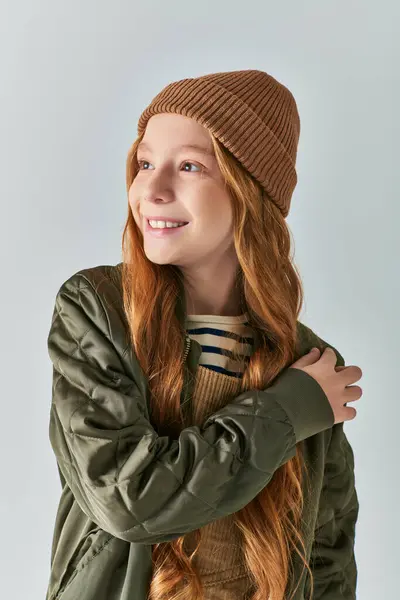 Зимняя мода, мечтательная девушка в вязаной шляпе и верхней одежде, смотрящая в сторону на сером фоне — стоковое фото