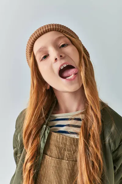 Preteen girl in knitted hat and outerwear sticking out tongue and looking at camera on grey backdrop — Stock Photo