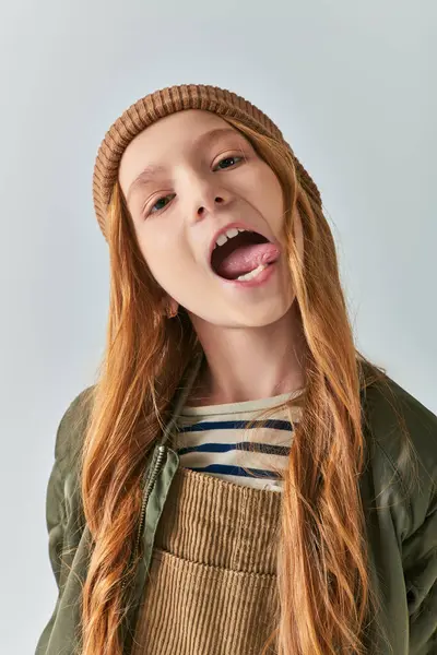 Preteen fille en bonnet tricoté et vêtements de dessus collant la langue et regardant la caméra sur fond gris — Photo de stock