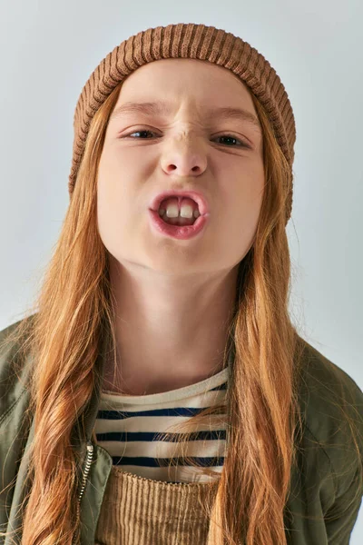 Angry preteen girl in knitted hat and outerwear showing teeth and looking at camera on grey backdrop — Stock Photo