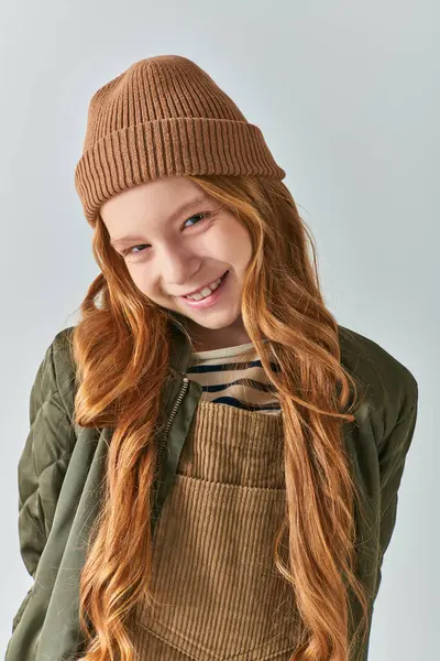 Stylish kid, cheerful girl in knitted hat and outerwear looking at camera on grey background — Stock Photo