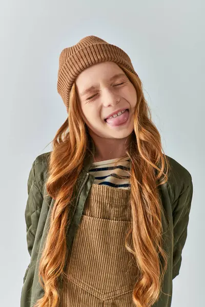 Niño con estilo, chica divertida en sombrero de punto y ropa de abrigo sobresaliendo lengua sobre fondo gris - foto de stock