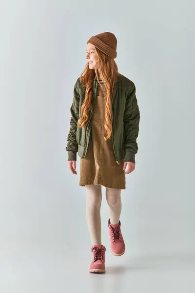 Niña preadolescente feliz con el pelo largo caminando en sombrero de invierno, chaqueta en la parte superior del vestido y botas en gris - foto de stock