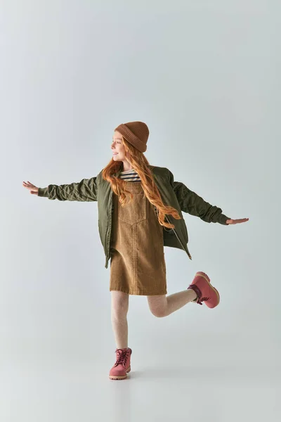 В полный рост взволнованная девушка в зимнем наряде и вязаной шляпе бегает с вытянутыми руками на сером — стоковое фото