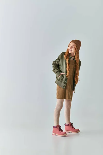 Comprimento total da menina feliz em roupa de inverno e chapéu de malha posando com a mão no bolso em cinza — Fotografia de Stock