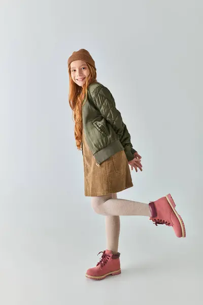 Full length of cheerful preteen girl in winter outfit with boots and hat looking at camera on grey — Stock Photo