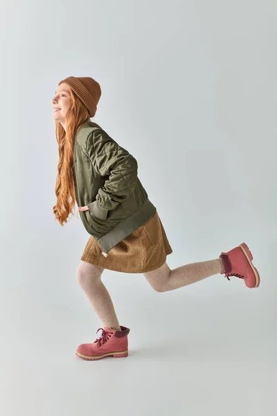 Longitud completa de la niña preadolescente juguetona en traje de invierno con botas y sombrero corriendo sobre fondo gris - foto de stock