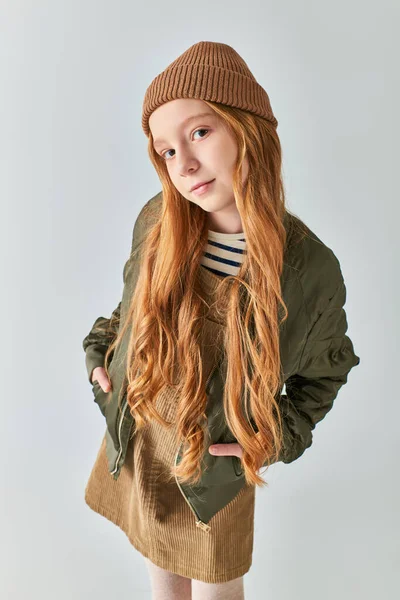 Preteen girl in winter outfit and hat posing with hands in pockets and looking at camera on grey — Stock Photo