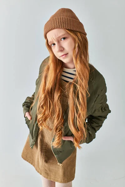 Das betroffene Mädchen im Winteroutfit und Hut posiert mit den Händen in den Taschen und auf grauem Hintergrund — Stockfoto