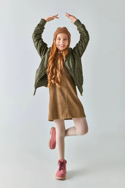 Веселая девушка в зимнем наряде в сапогах и шляпе, улыбающаяся и изображающая балерину на сером фоне — стоковое фото