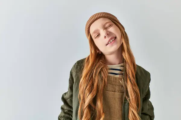 Cute girl in knitted hat grimacing and looking at camera on grey backdrop, children winter fashion — Stock Photo