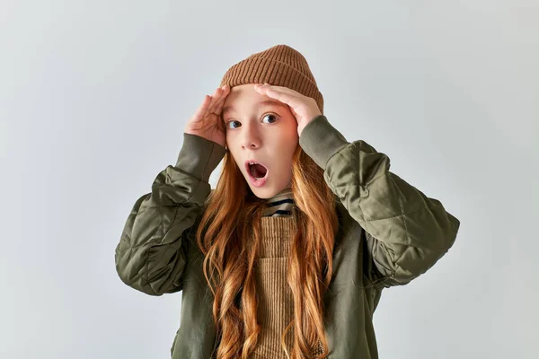 Expressive scared girl in stylish winter outfit with knitted hat puffing cheeks on grey backdrop — Stock Photo