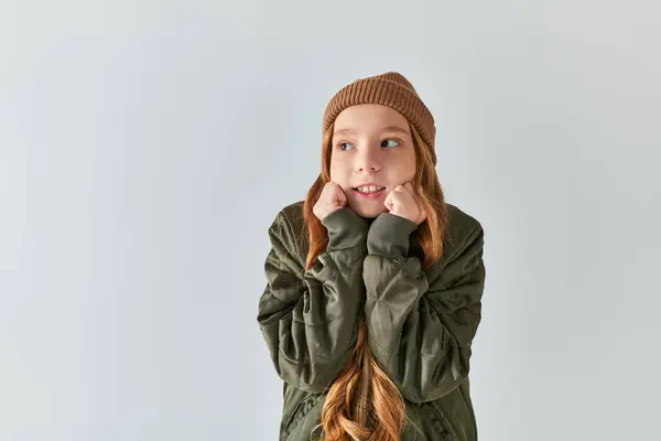 Preteen girl in stylish winter outfit with knitted hat feeling cold while standing on grey backdrop — Stock Photo