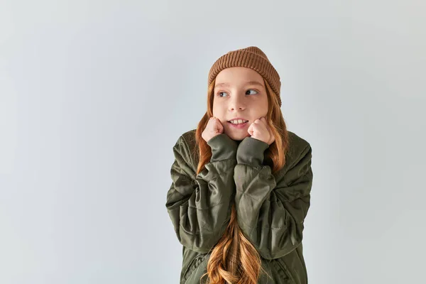 Preteen girl in stylish winter outfit with knitted hat feeling cold while standing on grey backdrop — Stock Photo