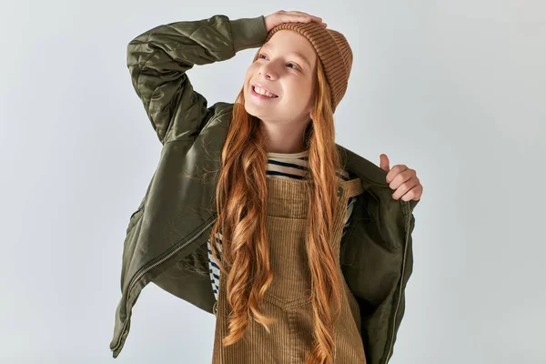 Довольная девочка-подросток в трикотажной шляпе и теплая одежда, регулирующая зимнюю куртку на сером фоне — стоковое фото