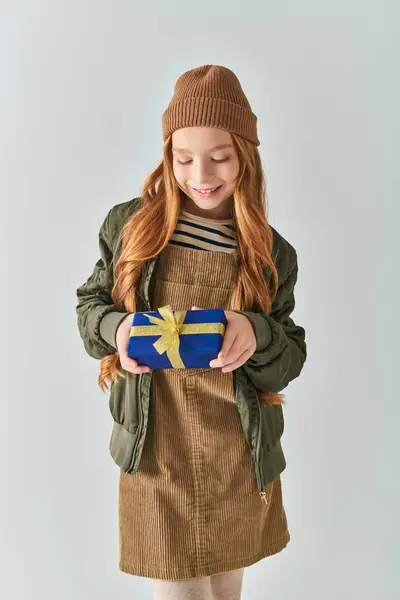 Fröhliches Mädchen in stylischem Winteroutfit mit Hut, der verpacktes Weihnachtsgeschenk auf grauem Hintergrund hält — Stockfoto