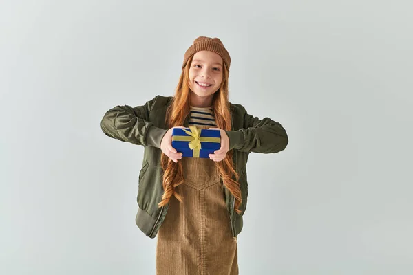 Menina feliz em roupa de inverno elegante com chapéu segurando embrulhado presente de Natal em pano de fundo cinza — Fotografia de Stock