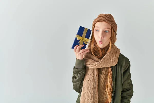 Preteen girl in stylish winter outfit with hat holding wrapped Christmas present, wow emotion — Stock Photo