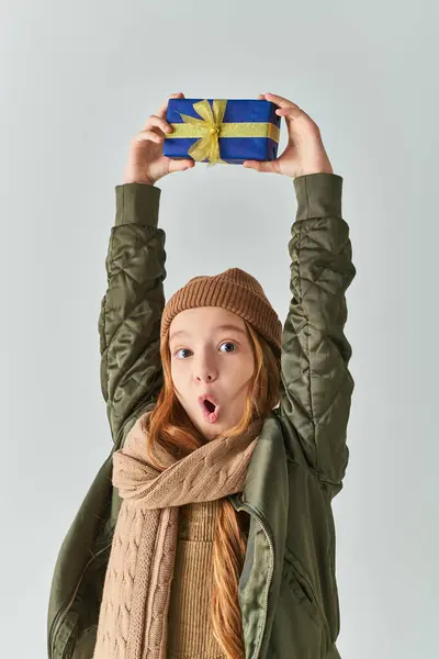 Amazed girl in stylish winter outfit with hat holding Christmas present above head on grey backdrop — Stock Photo