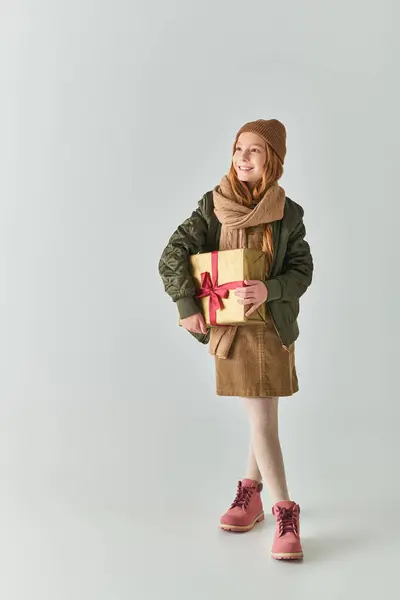 Comprimento total, menina feliz em roupa de inverno elegante com chapéu segurando presente de Natal no fundo cinza — Fotografia de Stock