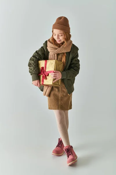 In voller Länge, fröhliches Mädchen im Winter-Outfit mit Hut hält Weihnachtsgeschenk auf grauem Hintergrund — Stockfoto