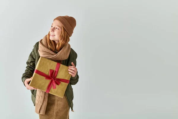 Lächelndes Mädchen im trendigen Winteroutfit mit Strickmütze hält Weihnachtsgeschenk vor grauem Hintergrund — Stockfoto