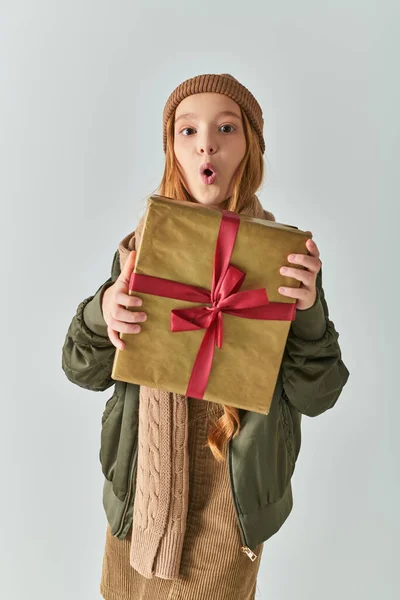 Amazed girl in warm clothes with winter hat holding Christmas present above head on grey backdrop — Stock Photo