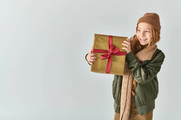 Niña preadolescente sonriente en traje de invierno con sombrero de punto con regalo de Navidad sobre fondo gris - foto de stock