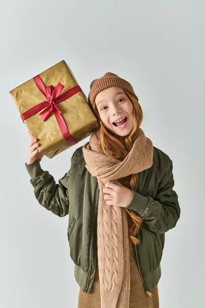 Niña preadolescente emocionada en traje de invierno con sombrero de punto con regalo de Navidad en el fondo gris - foto de stock