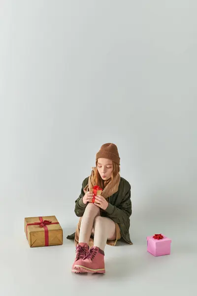 Niedliche preteen Mädchen im Winter-Outfit mit Strickmütze hält Weihnachtsgeschenk und sitzt auf grau — Stockfoto