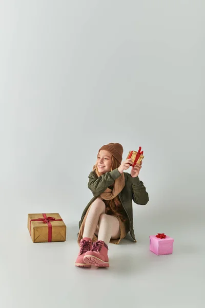 Happy preteen girl in winter outfit with knitted hat holding Christmas present and sitting on grey — Stock Photo