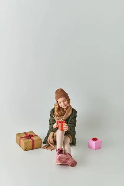 Fröhliches Mädchen im Winteroutfit mit Strickmütze, das ein Weihnachtsgeschenk in der Hand hält und auf grau sitzt — Stockfoto