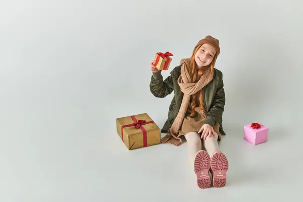 Happy preteen kid in stylish outfit with winter hat holding Christmas present and sitting on grey — Stock Photo