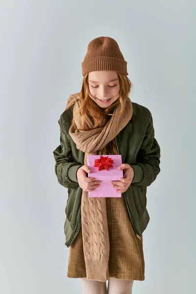Happy preteen kid in stylish outfit with winter hat holding Christmas present and standing on grey — Stock Photo
