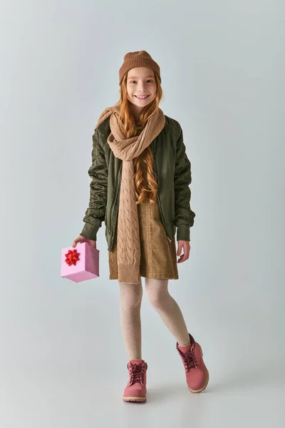 Fröhliches Mädchen in Winterkleidung und Strickmütze, das ein Weihnachtsgeschenk hält und auf grau steht — Stockfoto