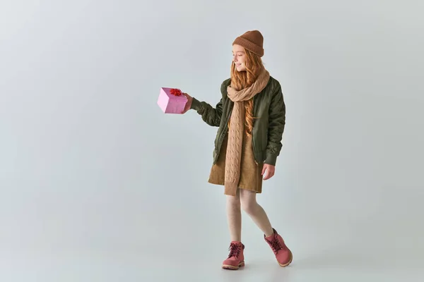 Menina pré-adolescente alegre em roupas de inverno e chapéu segurando presente de Natal e de pé no cinza — Fotografia de Stock