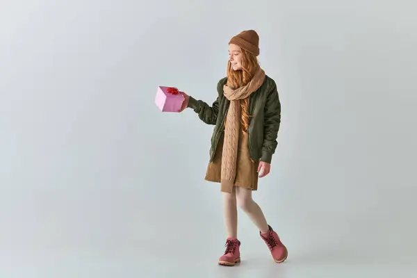 Fröhliche Preteen Mädchen in Winterkleidung und Hut mit Weihnachtsgeschenk und steht auf grau — Stockfoto