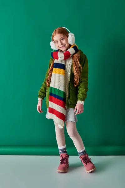 Capas acogedoras, chica feliz en orejeras y bufanda a rayas de pie en traje de invierno en turquesa - foto de stock
