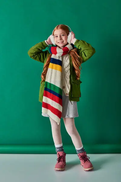 Couches confortables, fille gaie dans des cache-oreilles et foulard rayé debout en tenue d'hiver sur turquoise — Photo de stock