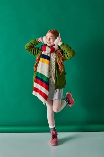 Camadas aconchegantes, menina alegre em regalos de orelha e cachecol listrado em pé na roupa de inverno em turquesa — Fotografia de Stock