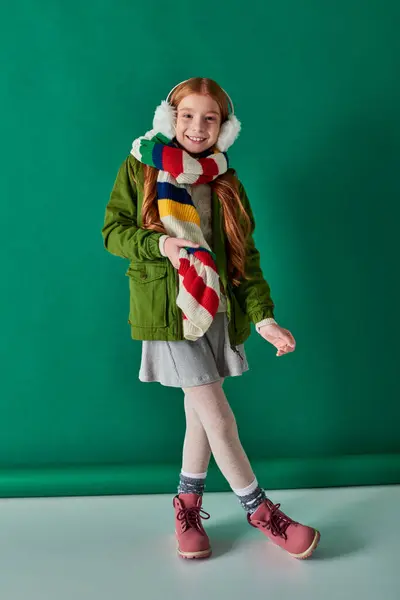 Chica feliz en orejeras y bufanda de pie en traje de invierno sobre fondo turquesa, capas acogedoras - foto de stock