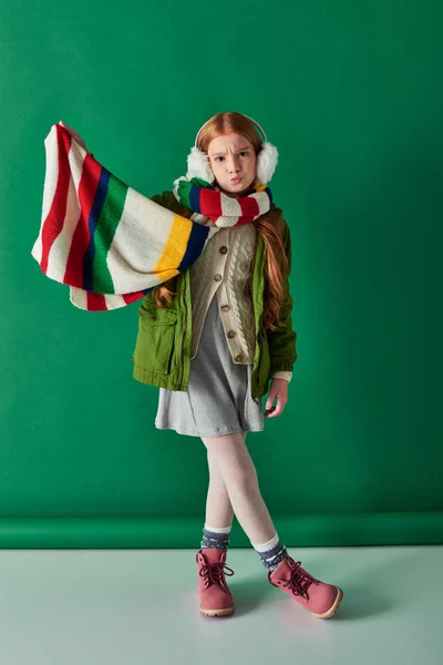 Criança pré-adolescente em abafadores de ouvido e cachecol em pé na roupa de inverno em pano de fundo turquesa, lábios amuados — Fotografia de Stock