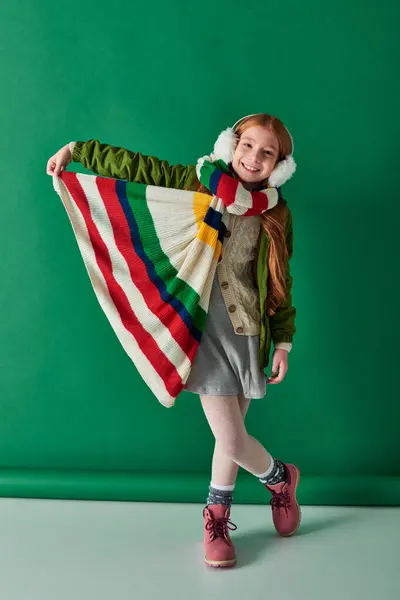 Enfant positif dans des cache-oreilles et écharpe debout en tenue d'hiver sur fond turquoise, couches confortables — Photo de stock