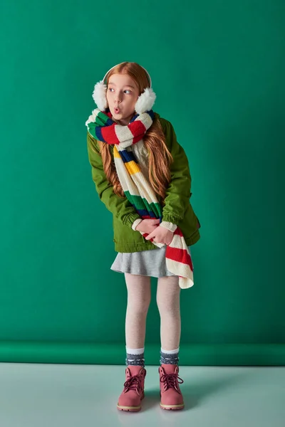 Pré-adolescente criança no ouvido regalos e cachecol de pé em roupas de inverno em pano de fundo turquesa, lábios amuados — Fotografia de Stock