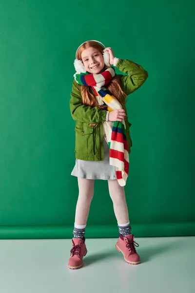 Niño positivo en orejeras y bufanda de pie en traje de invierno sobre fondo turquesa, capas acogedoras - foto de stock