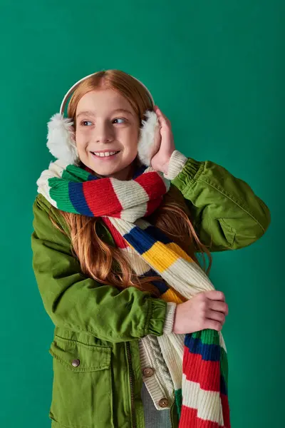 Niedliches Mädchen in gestreiftem Schal und Winterjacke, die Ohrenschützer auf türkisfarbenem Hintergrund berührt, gemütliche Schichten — Stockfoto